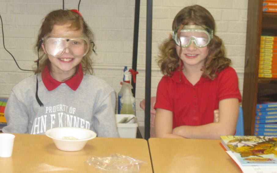 Two young girls in science goggles smiling at JFK Catholic School classroom.
