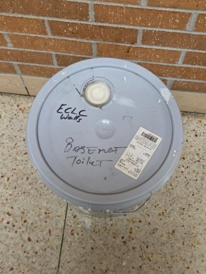 Trash bin labeled 'Basement Toilet' near brick wall at JFK Catholic School.